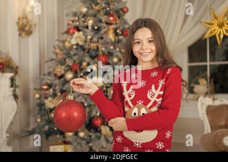 Le rouge est mon préféré. Décor d'attributs de Noël. Petite fille près de l'arbre de Noël. Enfant célébrer Noël à la maison. Traditions d'hiver. Boutique cadeaux concept. Adorable gamin excité porter le chandail de noël. Banque D'Images