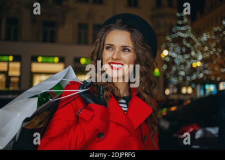 Hiver amusant. Heureuse élégante femme touristique solo en manteau rouge et béret noir avec des sacs de shopping à l'extérieur de la ville dans la soirée. Banque D'Images