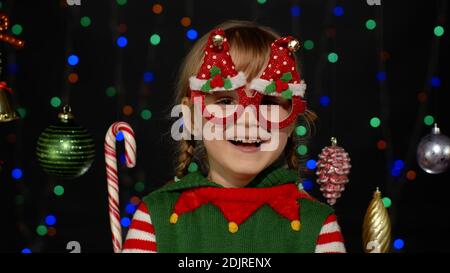 Petit blond enfant adolescent fille dans Noël elf Santa aide costume posant isolé sur fond noir. L'enfant se berce, faisant des visages amusants. Fête du nouvel an des gens Banque D'Images