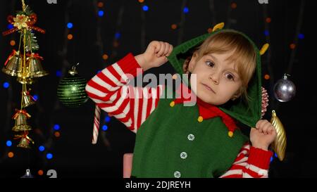 Petit blond enfant adolescent fille dans Noël elf Santa aide costume posant isolé sur fond noir. Enfant dansant, se berner, faire des visages drôles. Fête du nouvel an des gens Banque D'Images