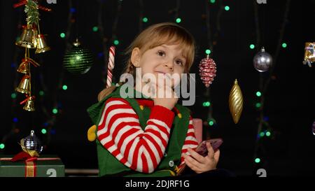 Enfant à la recherche de cadeaux, cadeau pour les amis et la famille, faire des achats en ligne en utilisant le téléphone mobile isolé sur le verrouillage de quarantaine de coronavirus. Enfant fille en costume d'aide de Noël elf Père Noël Banque D'Images