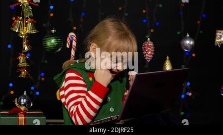 Enfant à la recherche de cadeaux, cadeau pour les amis et la famille, faire des achats en ligne en utilisant le téléphone mobile isolé sur le verrouillage de quarantaine de coronavirus. Enfant fille en costume d'aide de Noël elf Père Noël Banque D'Images