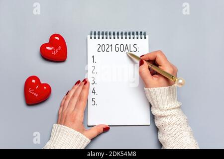 La main féminine tient le stylo et écrit 2021 buts du nouvel an sur le bloc-notes blanc, deux coeurs rouges sur fond gris vue du dessus Flat Lay motivation d'affaires, idée Banque D'Images