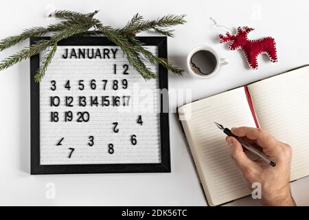 Main masculine avec un stylo et un carnet pour prendre des notes des buts et des plans pour la nouvelle année, calendrier et décoration d'arbre de Noël sur le bureau Banque D'Images