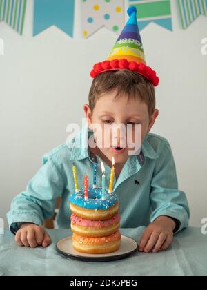 Un garçon de cinq ans souffle les bougies sur le gâteau de beignets de fête à son anniversaire. Petit enfant heureux et assiette avec beignets empiler le gâteau avec des bougies sur la table. Verticale. Banque D'Images