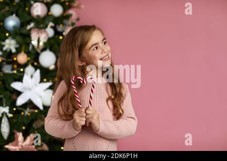 adorable petite fille joyeuse tenant une canne à sucettes en forme de coeur sur le fond rose de l'arbre de noël. espace de copie Banque D'Images