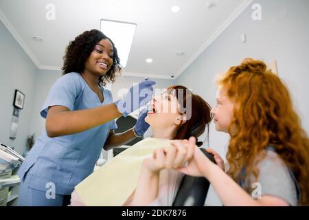 Vue latérale du bas de la jeune femme caucasienne ayant des dents de contrôle et de traitement. Sa petite fille tient la main de sa mère, tandis que la femme dentiste noir Banque D'Images