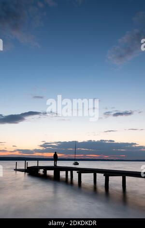 A single man in a hoodie stands on a wooden jetty on the banks of the Ammersee during sunset with a crescent moon. Stock Photo