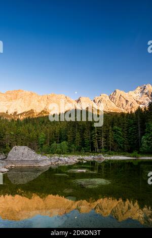 Reflet du massif du Wetterstein dans les Alpes allemandes, pendant la lumière du soir et trois quarts de lune dans un petit lac près de l'Eibsee. En arrière-plan le Zugspitze. Banque D'Images