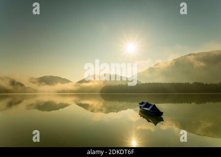 Un seul bateau à rames avec bâche est amarré au lac Sylvensteinspeicher, tandis que le soleil envoie ses rayons au-dessus du lac avec un léger brouillard tôt le matin et se reflète dans l'eau. Banque D'Images