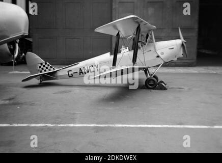 Une photographie vintage en noir et blanc de 1954 montrant un avion de Havilland Tiger Moth, enregistrement G-ANCY, garé à l'extérieur d'un hangar. Banque D'Images