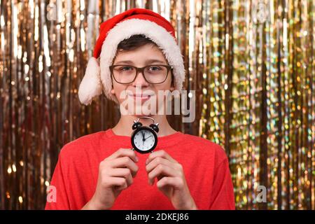 Portrait d'un garçon avec des lunettes et dans un chapeau de Père Noël avec une horloge indiquant l'heure sur un fond doré. Décoration de Noël. Douze heures Banque D'Images