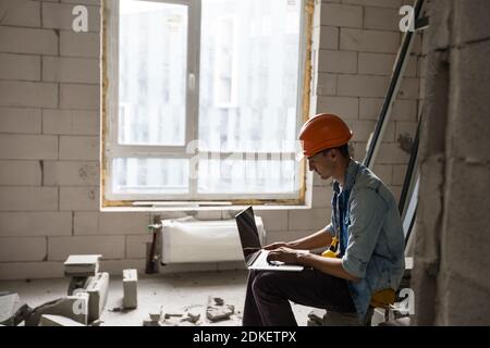 Beau architecte ou contremaître dans un casque travaillant avec un ordinateur portable les dessins d'appartement à la structure intérieure blanche Banque D'Images