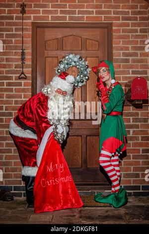 Royaume-Uni, Quorn - décembre 2020 : le père Noël et un Elf se faufilent devant une porte, car ils livrent des cadeaux d'un grand sac rouge Banque D'Images