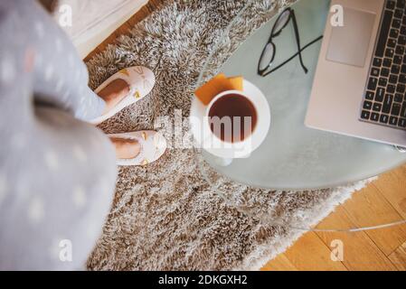 Les pieds et les jambes d'une jeune femme portant une tenue décontractée vêtements de maison ou de pyjama et chaussons de chatte rose et debout sur la moquette près de la table basse avec du café Banque D'Images