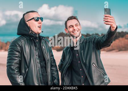 Two Handsome Male Friends Making Selfie Using Black Smartphone at the Beach. Youthful Men in Black Clothes Having Fun by Making Photos Against the Sea Banque D'Images