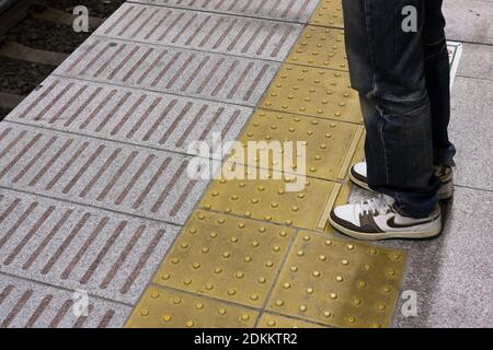 Jambes d'un passager attendant derrière une ligne de sécurité jaune faite de blocs Tenji pour l'utilisation par les aveugles ou les malvoyants. À la gare de Yokohama, Japon. Banque D'Images