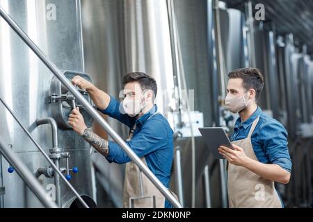 Production de bière à l'usine pendant l'épidémie de Covid-19. Les employés travaillant sur des tabliers et des masques de protection avec une tablette numérique travaillent à la main avec l'équipement et les contrôles Banque D'Images