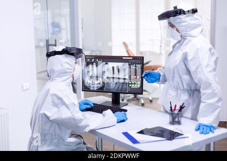 Dentiste et infirmière discutant des problèmes de dents du patient vêtus d'un équipement de protection individuelle. Spécialiste médical portant un équipement de protection contre le coronavirus lors d'une épidémie mondiale qui regarde la radiographie dans un cabinet dentaire. Banque D'Images