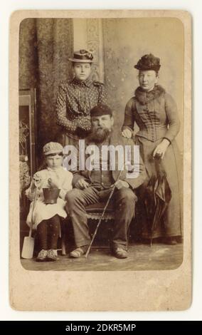 Original de caractère victorien CDV (carte de visite) photographie souvenir d'une famille victorienne de classe ouvrière avec belle-mère en vacances à la station balnéaire de Skegness, Lincolnshire, Angleterre, Royaume-Uni vers 1888. La mère est attirante. Père est barbu. L'enfant tenant un seau et une bêche porte un curieux mélange de robe de fille et de casquette de garçon et, étant donné l'habitude victorienne d'habiller les garçons dans des vêtements de fille, est probablement un garçon. Il n'a pas l'air très heureux et se fait apparemment prendre en photo sous la contrainte. Banque D'Images