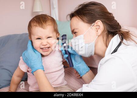 Le pédiatre examine l'oreille de la petite fille à la maison pendant la quarantaine pandémique COVID-19 du coronavirus. Médecin utilisant l'otoscope (auriscope) pour vérifier le canal auditif a Banque D'Images