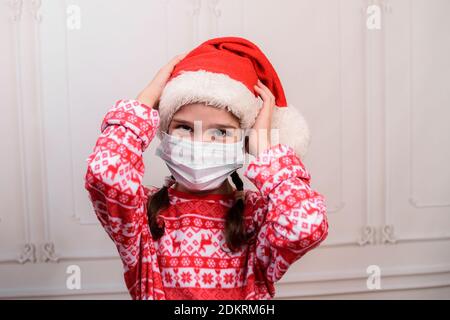 Une petite fille amusante portant un chapeau de père Noël et un masque médical. Enfant fille à expression heureuse le jour de noël. Banque D'Images