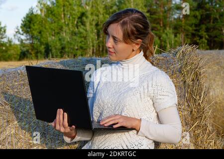 adorable jeune fille dans un chandail blanc sur fond de botte de foin et de forêt verte tient un ordinateur portable dans elle mains Banque D'Images