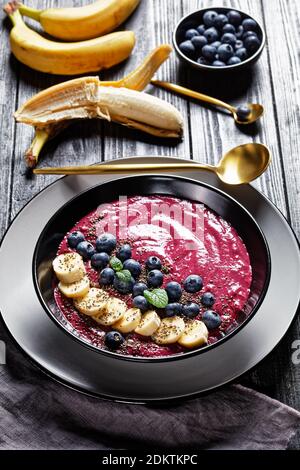 Bol à smoothie aux myrtilles et aux bananes avec yaourt, noix de cajou, graines de chia et graines de lin sur une assiette noire sur fond de bois rustique foncé avec ingrédient Banque D'Images