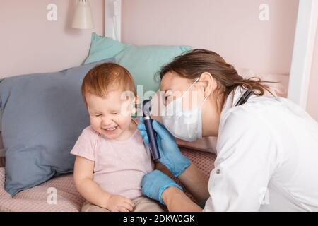 Le pédiatre examine l'oreille de la petite fille à la maison pendant la quarantaine pandémique COVID-19 du coronavirus. Médecin utilisant l'otoscope (auriscope) pour vérifier le canal auditif a Banque D'Images