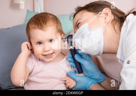 Le pédiatre examine l'oreille de la petite fille à la maison pendant la quarantaine pandémique COVID-19 du coronavirus. Médecin utilisant l'otoscope (auriscope) pour vérifier le canal auditif a Banque D'Images