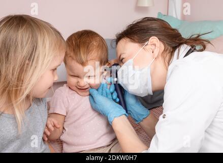 Le pédiatre examine l'oreille de la petite fille à la maison pendant la quarantaine pandémique COVID-19 du coronavirus. Médecin utilisant l'otoscope (auriscope) pour vérifier le canal auditif a Banque D'Images