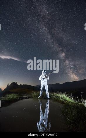 Cosmonaute dans l'espace costume jouant de la guitare sous le ciel bleu de nuit avec la voie lactée. Guitariste se reflétant dans un petit lac tout en se tenant dans une vallée herbeuse illuminée. Concept de musique, de cosmonautics et de galaxie. Banque D'Images