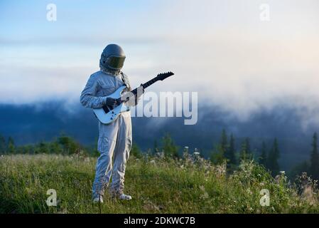 Spaceman en costume blanc et casque tenant une guitare blanche debout sur un magnifique pré de montagne verte le matin, le brouillard monte du val Banque D'Images