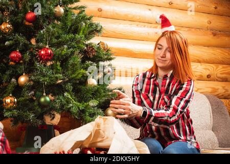 Une femme gaie dans une chemise à carreaux pend une belle boule brillante sur un arbre de Noël sur le fond d'une cheminée éclairée, des chaussettes suspendues pour des cadeaux. Banque D'Images