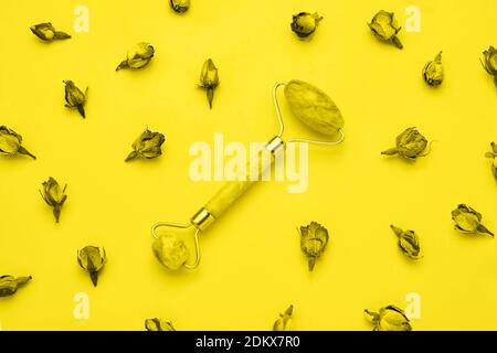 Rouleau de massage pour le visage avec deux pierres de jade sur fond jaune avec des fleurs de roses séchées autour. Concept de soins de santé. Vue de dessus Banque D'Images