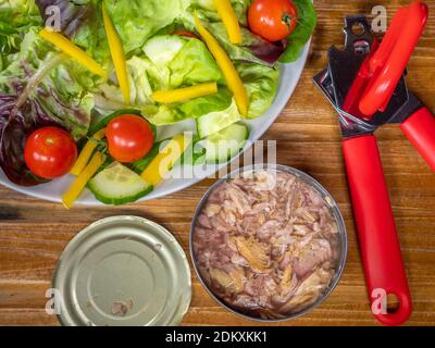 Gros plan POV d'un ouvre-boîte rouge à côté d'une boîte de thon rouge haché dans l'eau, avec le couvercle enlevé, à côté d'une assiette de salade. Banque D'Images