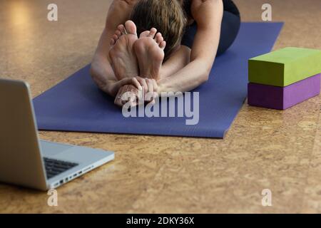 Femme pratique paschimottanasana C yoga pose devant l'ordinateur portable avec des accessoires par tapis. Yogi femelle faisant un coude vers l'avant assis. Cours en ligne, bien-être Banque D'Images