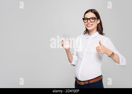 souriante jeune femme d'affaires enceinte dans des verres avec un verre d'eau le pouce vers le haut est isolé sur le gris Banque D'Images