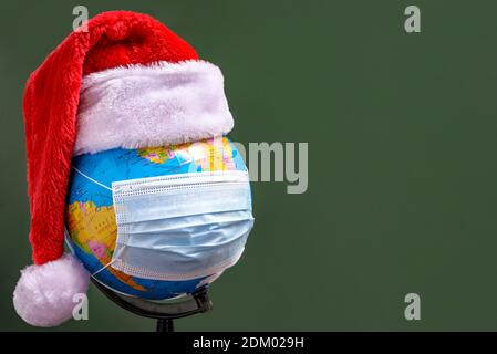 Earth globe with Santa Claus hat and blue surgical face mask at green background. The concept of celebrating the New Year and Christmas during the COV Stock Photo