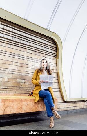 une femme heureuse en tenue d'automne souriant à l'appareil photo tout en étant assise avec journal sur plate-forme de métro Banque D'Images