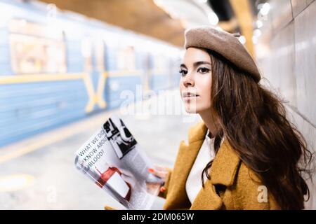 jeune femme dans le magazine beret holding en étant assise dans le métro plate-forme avec train flou Banque D'Images