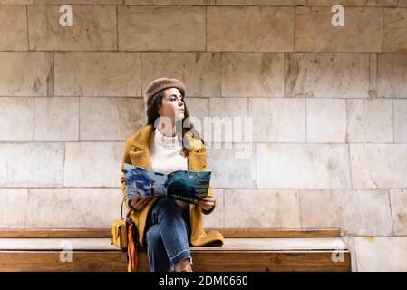 femme élégante avec tenue automnale, avec un regard à l'extérieur tout en tenant le magazine sur le banc de plate-forme de métro Banque D'Images