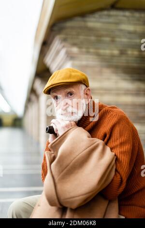 homme âgé en casquette et chandail regardant loin en étant assis avec un bâton de marche sur la plate-forme de métro Banque D'Images