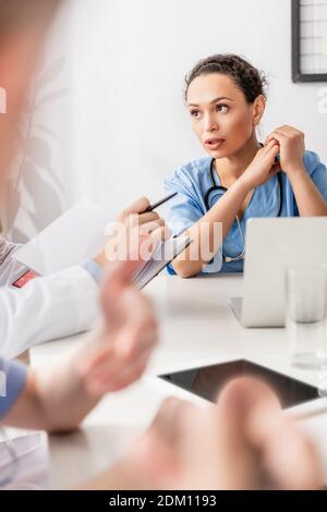 Infirmière afro-américaine regardant un collègue avec un ordinateur portable à proximité d'appareils sur un premier plan flou de la table Banque D'Images