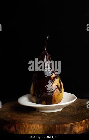 Une poire cuite au sirop de chocolat sur une assiette blanche sur fond sombre Banque D'Images