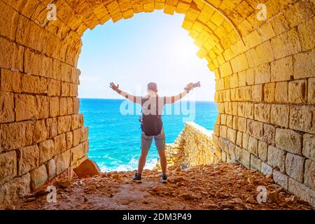 Guy voyageur avec sac à dos et mains levées, tenant l'appareil photo se tient sur la falaise dans une ancienne arche de pierre contre le fond de l'océan Banque D'Images