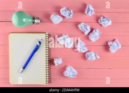 Notepad with a pen, incandescent bulb and crumpled white paper balls on a pink wooden table. Ideological concept, top view Stock Photo