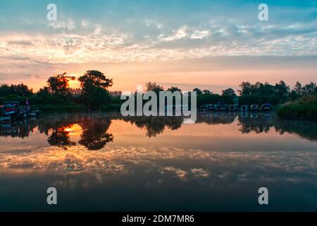 L'aube et le lever du soleil au-dessus d'une marina en été avec les couleurs du ciel reflétées dans le lac entouré de bateaux et d'arbres. Banque D'Images