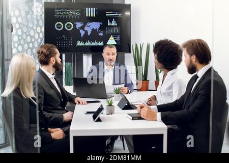 Homme d'affaires caucasien mature fait rapport à ses collègues multiethniques divers dans la salle de conférence, avec carte du monde, gapigraphics, graphiques Banque D'Images