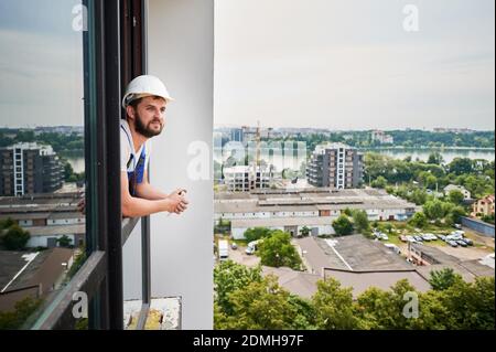 Jeune homme barbu dans un casque de sécurité profitant de la vue sur la ville tout en s'inclinant par la fenêtre de l'appartement. Plombier mâle regardant la rue avec des bâtiments, des arbres et de la rivière. Concept de vie urbaine, bâtiment Banque D'Images